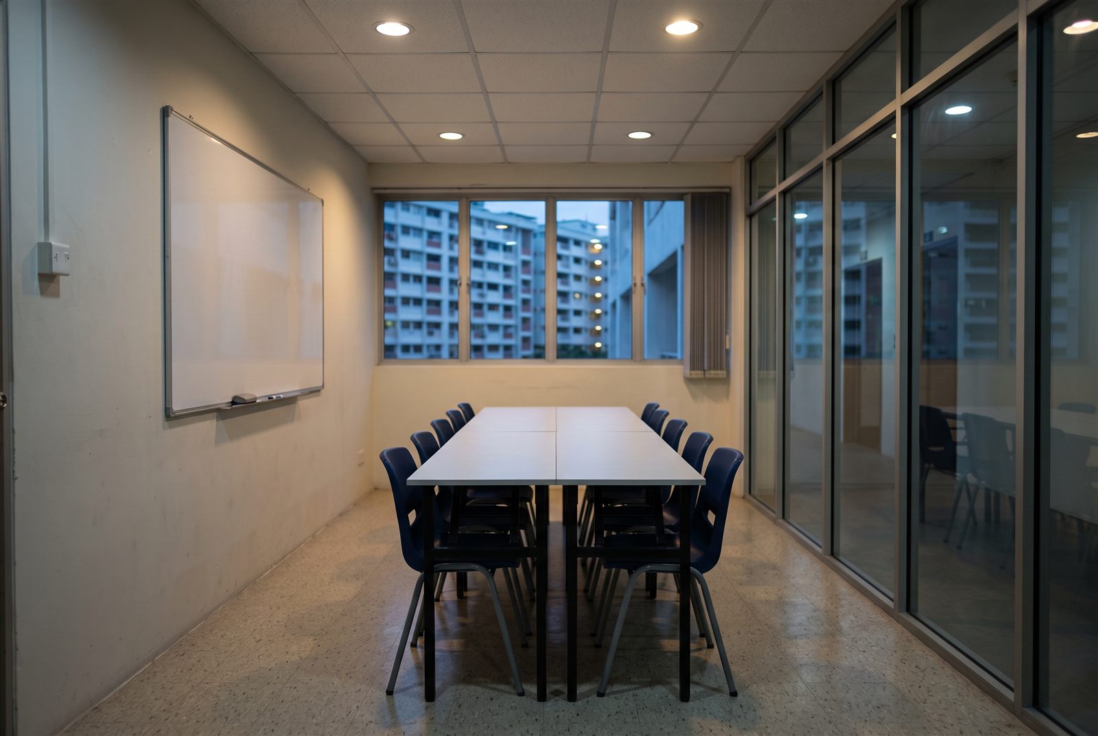An empty tuition classroom in the early evening: a long white table, a row of empty navy chairs, a blank whiteboard and HDB flats through the window.